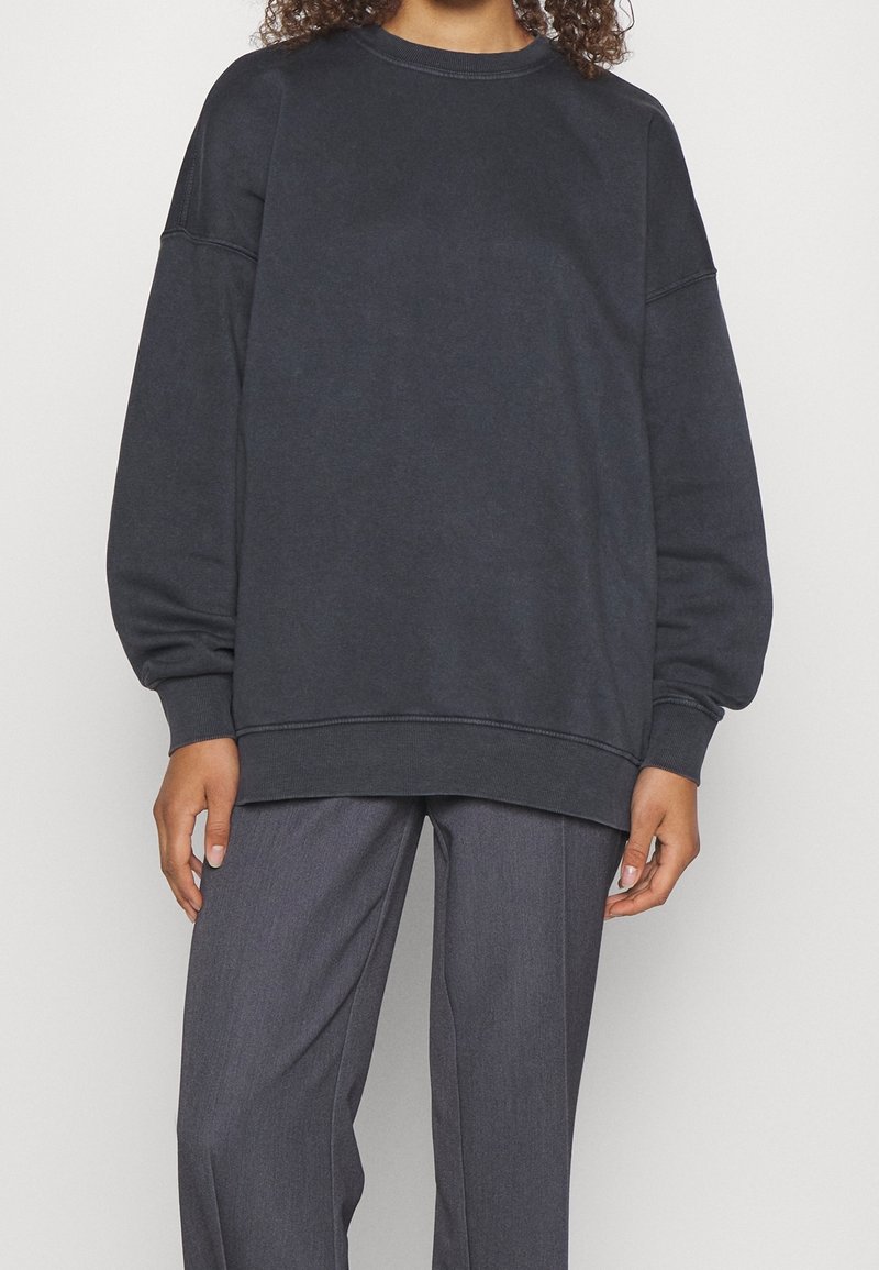 Sweatshirt oversized grigio scuro realizzato in tessuto morbido, con spalle scese e scollatura rotonda, abbinato a pantaloni grigi sartoriali.