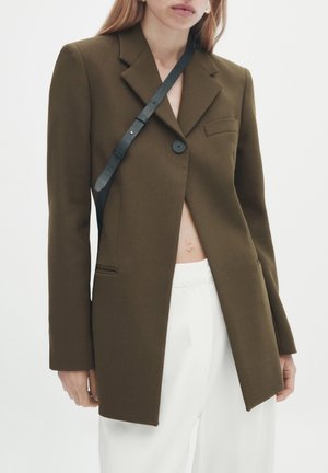 Vrouw draagt een olijfgroene colbert met één knoop over witte broek en een zwarte crossbodyriem tegen een effen lichte achtergrond.