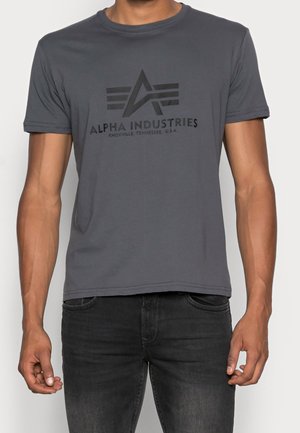 Mand iført en tætsiddende grå Alpha Industries t-shirt med sort logo og tekst, kombineret med mørkeblå jeans, stående foran en ensfarvet baggrund.