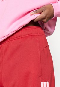 Piros sportnadrág texturált felülettel, elastikus derékkal, varrás részletekkel és egy oldalsó zsebbel. Fehér csíkos díszítéssel az alján.