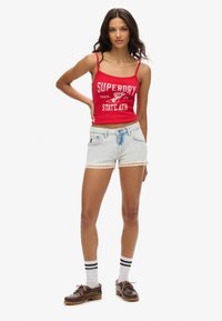 Top corto rojo con el texto "SUPERDRY", pantalones cortos de mezclilla azul claro con borde de encaje, calcetines blancos con rayas negras y zapatos marrones.