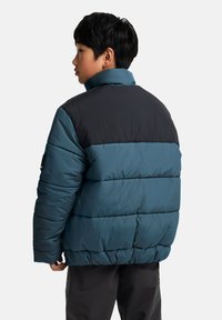 Teal og svart polstret jakke, med høy krage, horisontale sømmer og avslappet passform. Jevn tekstur med en myk finish.