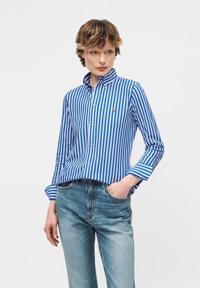 Άτομο που φοράει μπλε και λευκό κάθετο ριγέ πουκάμισο με κουμπιά και ανοιχτόχρωμο μπλε τζιν, με το ένα χέρι στην τσέπη, κοιτάζοντας πλάγια.