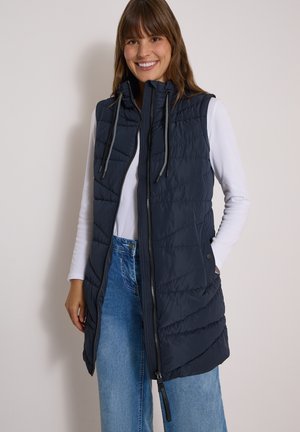 Kvinde smiler iført en sort quiltet vest uden ærmer over en hvid langærmet skjorte og blå jeans mod en ensartet lys baggrund.