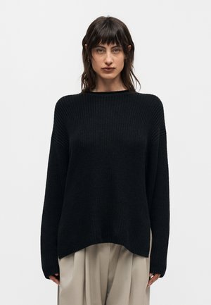 Žena s tamnom kosom odjevena u crni pleteni džemper i bež široke hlače stoji ispred jednobojne svijetle pozadine.