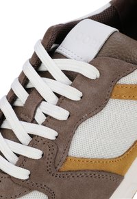 Sneaker in suede e pelle marrone con pannelli in rete bianca, lacci bianchi e una striscia di accento gialla; presenta una lingua testurizzata e un dettaglio del logo.