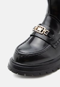 Mocassin en cuir verni noir avec semelle épaisse et détail de chaîne décoratif doré sur le devant.