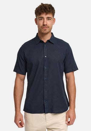 Ung mand med kort brunt hår iført en mørk marineblå kortærmet skjorte og lyse beige bukser mod en ensfarvet baggrund.