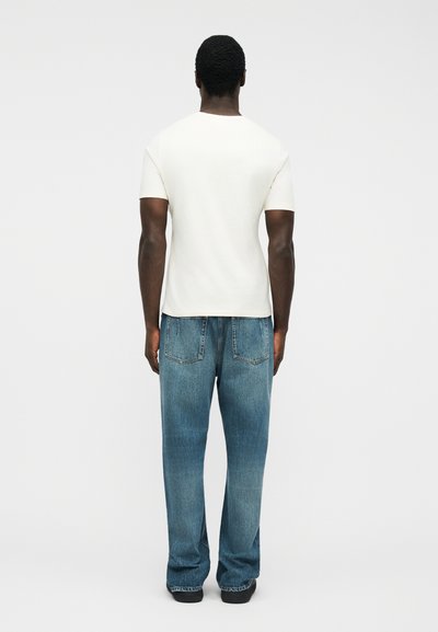 Άνδρας που στέκεται με την πλάτη προς τα εμπρός, φοράει απλό άσπρο κοντομάνικο πουκάμισο και χαλαρά μπλε τζιν παντελόνια, πάνω σε λευκό φόντο.