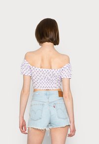 Top crop branco com ombros à mostra e padrão floral roxo, combinado com calções de ganga de lavagem clara com bainha desfiada e bolsos traseiros.