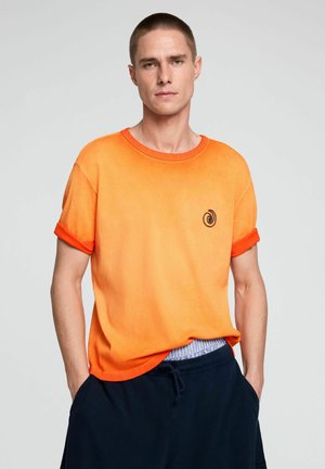 Junger Mann mit kurzen Haaren, der ein orangefarbenes T-Shirt mit Farbverlauf und hochgekrempelten Ärmeln sowie marineblaue Hose mit Kordelzug trägt, steht mit den Händen in den Taschen.