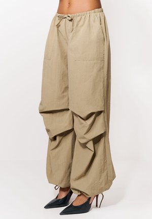 Beżowe szerokie spodnie z ściągaczami ze sznurkami w talii i na dole nogawek, noszone z czarnymi szpilkami o zaostrzonych noskach.