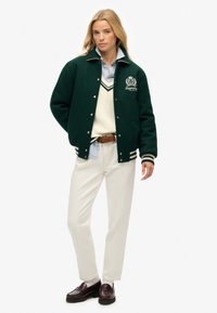 Groene wollen varsityjack met witte accenten, geborduurd logo, over een crèmekleurige trui en een lichtblauw gestreefd shirt, gecombineerd met witte broek.