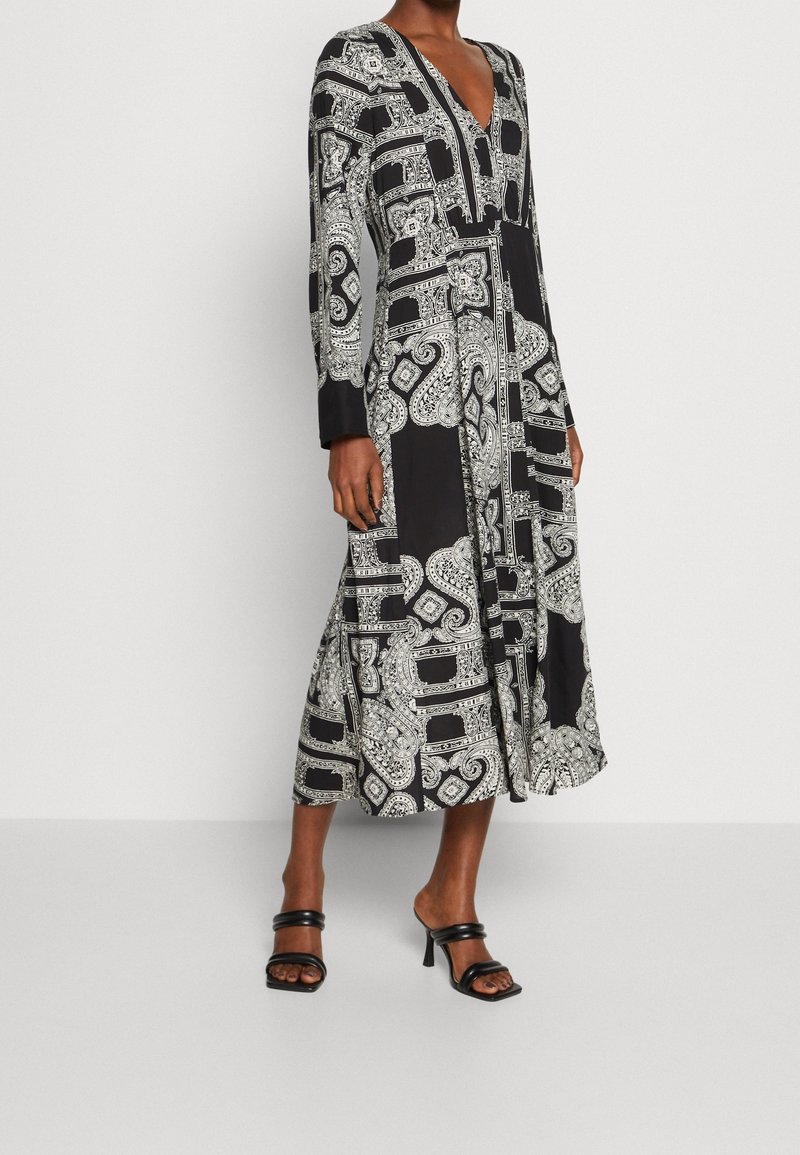 Robe noire et blanche à manches longues avec un décolleté en V, des motifs paisley complexes et une silhouette fluide qui arrive mi-mollet.