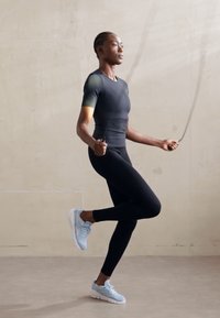 Sort sports skjorte med korte ærmer, sorte leggings, lyseblå sneakers; person der hopper på sjippetov, glat baggrund med subtil tekstur.