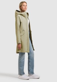 Femme portant un manteau à capuche vert clair, une chemise blanche, un jean bleu et des baskets blanches, debout sur un fond uni, de profil.