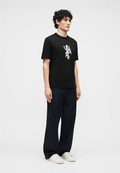 Νεαρός με σγουρά μαλλιά που φοράει μαύρο T-shirt με γραφικό αστροναύτη, σκούρο παντελόνι με ριγέ ευρύ πόδι και λευκά αθλητικά παπούτσια, στέκεται ακίνητος.