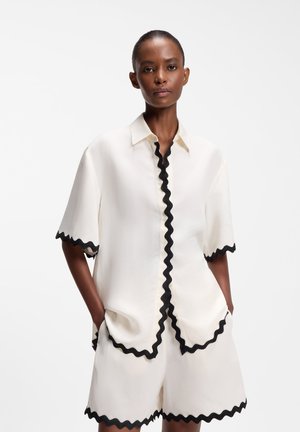 Kobieta ubrana w biały zestaw z krótkim rękawem i szortami z czarną falbanką, ręce w kieszeniach, stoi na białym tle.