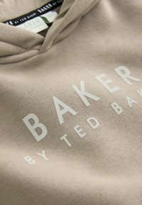 Bež hoodie napravljen od mekanog materijala s velikim bijelim logotipom "BAKER" i manjim natpisom "by TED BAKER" koji je istaknuto prikazan na prednjoj strani.
