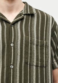 Κοντινό πλάνο ενός ατόμου που φοράει πουκάμισο ελιάς με λευκές κάθετες ρίγες, μπροστινή τσέπη και γιακά, κουμπωμένο μπροστά.