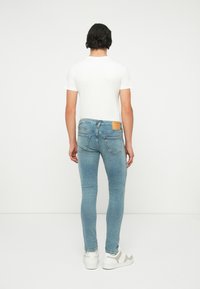 Jeans skinny azzurri chiari con tasche posteriori, indossati con una t-shirt bianca semplice e sneakers bianche. Presentano un'etichetta in pelle marrone chiaro.