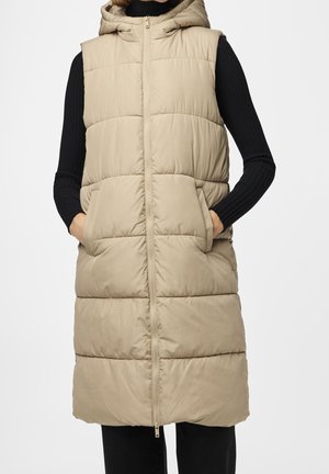 Beige gewatteerd mouwloos lang gilet met capuchon en rits, gedragen over een zwarte geribbelde top met lange mouwen, handen in de zakken, staand tegen een effen achtergrond.