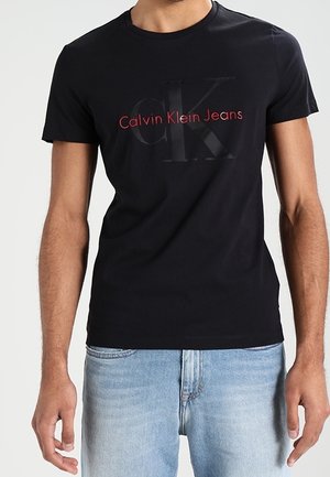 Mężczyzna noszący czarną koszulkę Calvin Klein Jeans z czerwono-czarnym logo, w połączeniu z jasnoniebieskimi jeansami z denimu.
