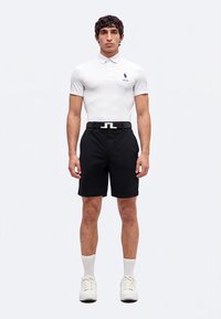 Homme debout portant un polo blanc, un short noir, des chaussettes blanches, des baskets blanches et une ceinture noire avec une boucle argentée.