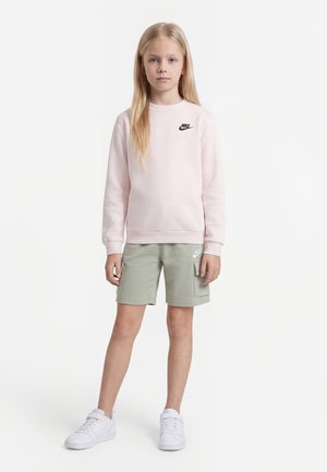Jeune fille debout, portant un sweat-shirt Nike rose pâle, un short cargo vert clair et des baskets blanches, sur un fond blanc uni.