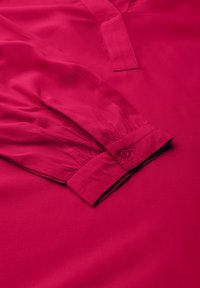 Blusa fucsia con escote en V, mangas largas y puños abotonados; textura suave con ligero brillo y detalles de costura sutiles.