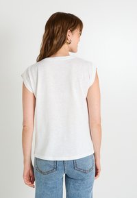 Camiseta blanca de manga corta con cuello redondo y puños enrollados, hecha de un tejido ligero. Llevada con jeans de cintura alta en azul.