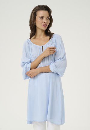 Lichtblauwe tuniek met lange mouwen, een ronde halslijn en geplooide textuur. Voorzien van een knoopsluiting op de borst en een ontspannen silhouet.