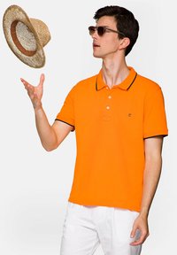 Jasnopomarańczowa koszulka polo z czarnymi akcentami, krótki rękaw, trzy guziki przy dekolcie. Model w okularach przeciwsłonecznych, rzucający słomkowy kapelusz.