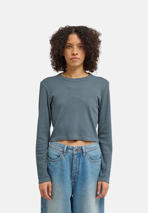 Persona con capelli ricci indossa una maglia blu corta a maniche lunghe con la scritta ""element"" e jeans blu larghi, in piedi davanti a uno sfondo semplice.