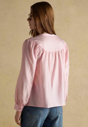 Woman with long brown hair and glasses wearing a loose light pink blouse with gathered yoke and blue jeans, facing away against a beige background.