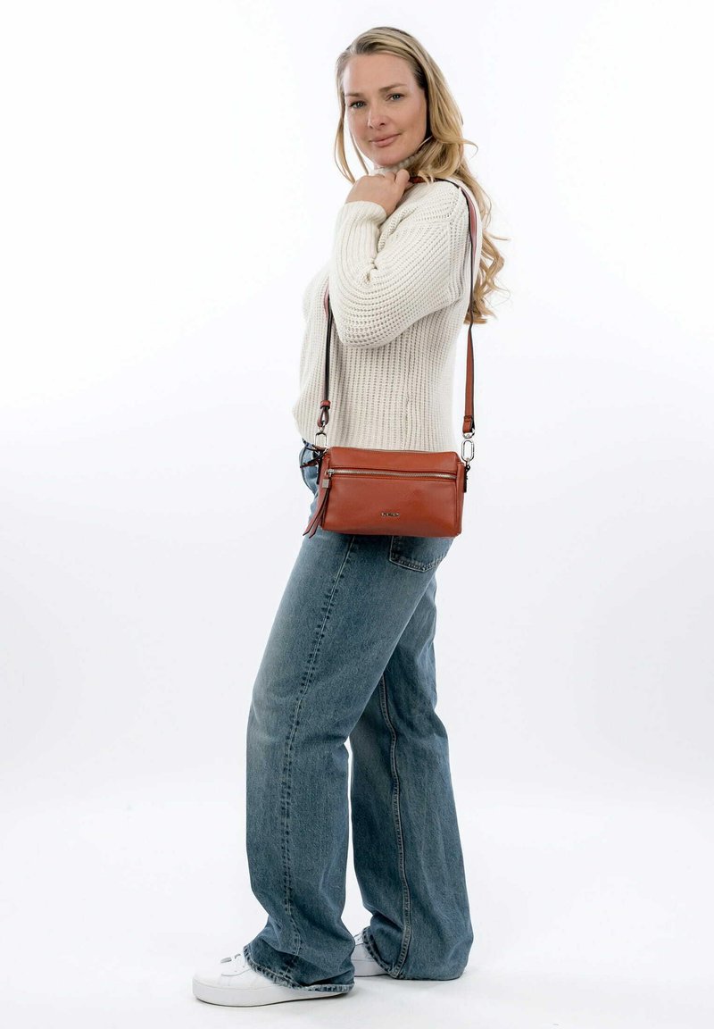 Borsa a tracolla in pelle marrone con tasca con zip, texture liscia e tracolla regolabile, indossata da una persona con un maglione chiaro e jeans.