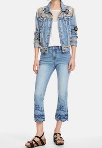 Chaleco de mezclilla con bordado floral en los hombros, top de encaje blanco, jeans de longitud tobillo en azul claro con dobladillo de patrón azul y sandalias negras.