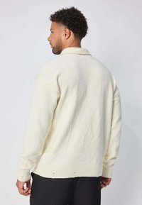 Pull en polaire crème avec des poignet et un ourlet côtelés, offrant une coupe décontractée et un col. La surface texturée ajoute de la profondeur.