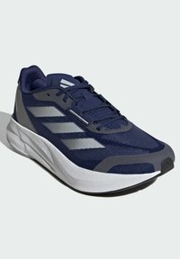 Scarpa da ginnastica navy con tomaia in rete, accenti grigi e tre strisce riflettenti. Suola leggera con texture distintiva e design della punta arrotondata.