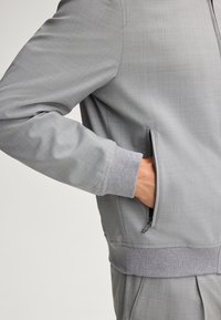 Graue, leichte Jacke mit strukturiertem Finish, ausgestattet mit einer seitlichen Zipptasche und gerippten Bündchen. Das Design zeichnet sich durch eine einfarbige Gestaltung und schlichte Linien aus.