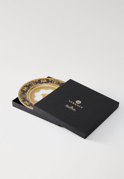 Dekoratívny tanier Versace v čiernej krabičke, zdobený zložitými zlatými vzormi na bielom pozadí s centrálnym motívom a značkovými detailmi.