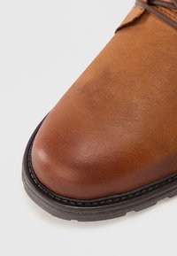 Ruskea nahkakenkä, jossa on pyöreä kärki, sileä pinta, musta kumipohja ja näkyvät ompeleet reunoilla.