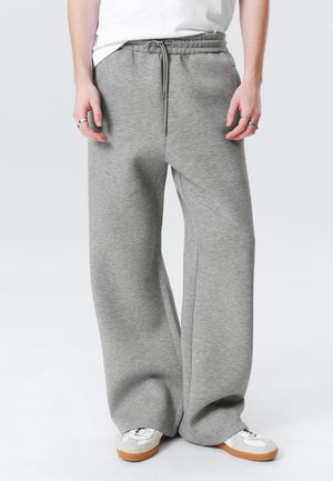Person, der weite graue Jogginghosen und weiße Sneakers mit Gummisohlen trägt, steht vor einem hellen Hintergrund.