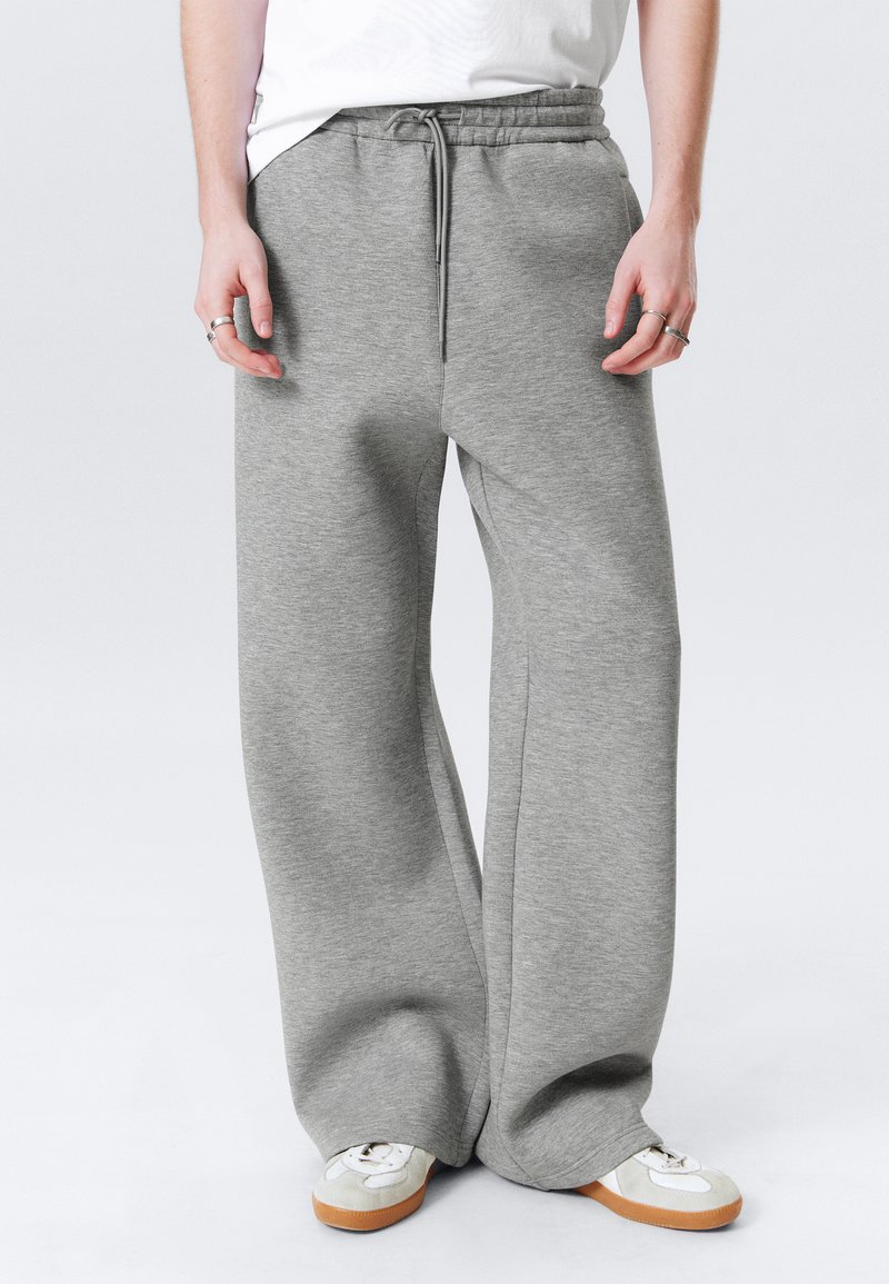 Person kledd i løse grå joggebukser og hvite sneakers med gummisåler, stående mot en lys bakgrunn.