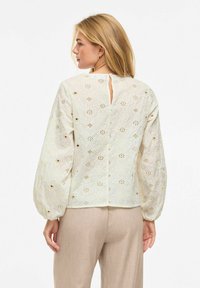 Femme aux cheveux blonds portant un chemisier ivoire en dentelle avec des découpes florales et un pantalon beige, vue de dos sur un fond uni.