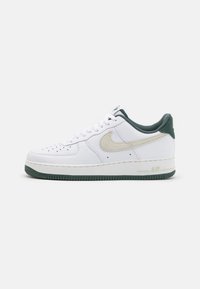 Bílé tenisky Nike Air Force 1 s texturovaným koženým svrškem, zelenými akcenty na patě a podrážce a béžovým logem Swoosh.