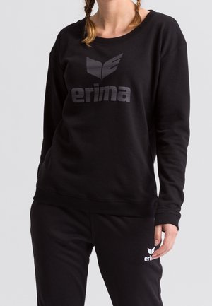 Personne portant un sweat-shirt et un pantalon noirs avec des logos Erima gris et blancs sur la poitrine et la cuisse, sur un fond uni.