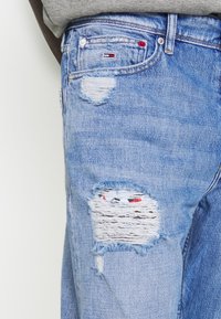 Světle modré džíny vyrobené z denimu, s poškozenými místy, třepenými okraji, malou logovou nálepkou a tradičním pěti-pocket designem.