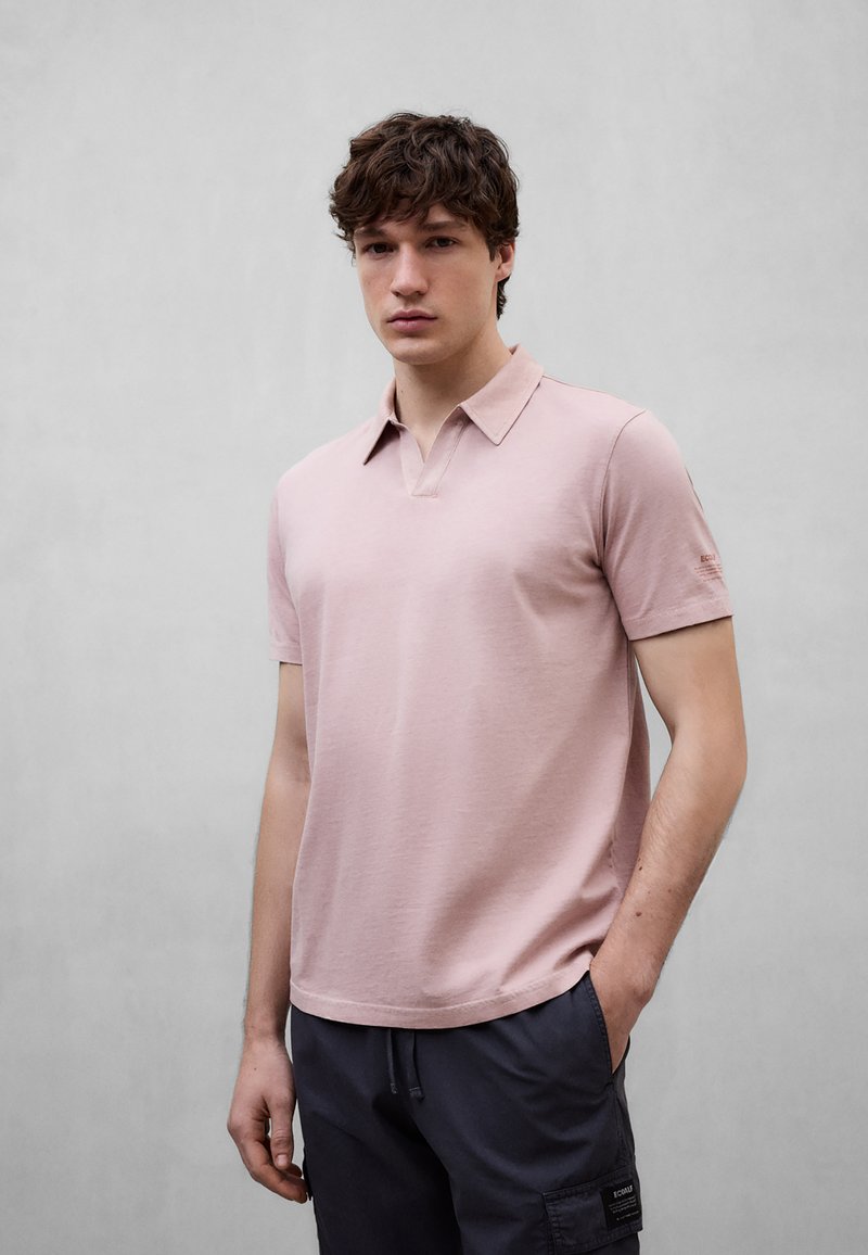 Jeune homme avec la main dans la poche portant un polo rose clair à manches courtes et un pantalon cargo foncé sur un fond gris uni.
