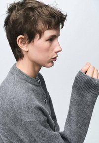 Suéter gris de punto con cuello alto, que presenta grandes botones de metal, mangas largas y puños acanalados. Textura suave, diseño minimalista.